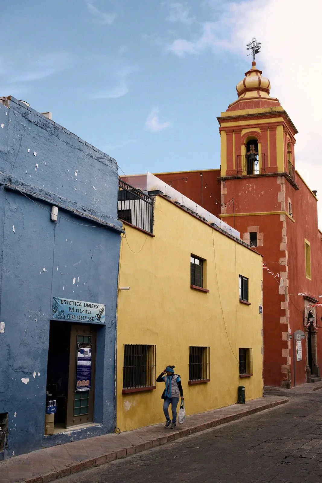 WorthWait: Fotoğrafları Görmek İçin 24 Saat Bekleten Kamera Uygulaması 21 A person walks along a cobblestone street past a blue building, a yellow building, and a tall, red church tower under a blue sky.