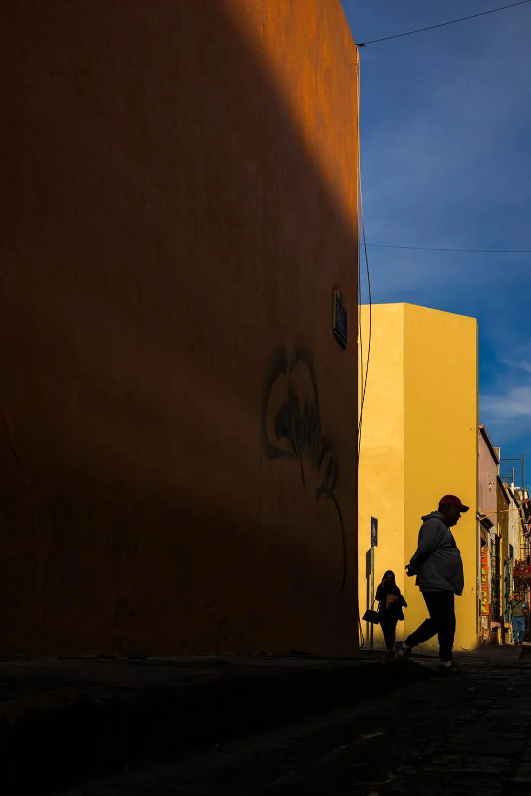 WorthWait: Fotoğrafları Görmek İçin 24 Saat Bekleten Kamera Uygulaması 22 A person in a red cap and grey hoodie walks along a street past a large, shadowed orange wall, with a second person walking behind them.