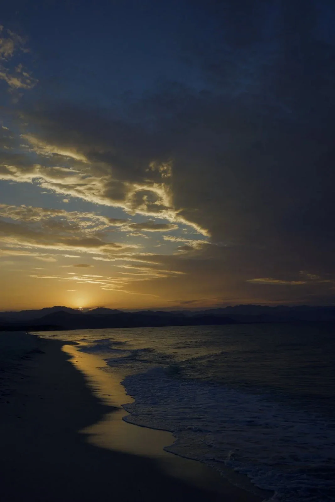 WorthWait: Fotoğrafları Görmek İçin 24 Saat Bekleten Kamera Uygulaması 23 A sunset over a calm ocean with waves washing onto a sandy beach, framed by dark clouds and distant mountains.