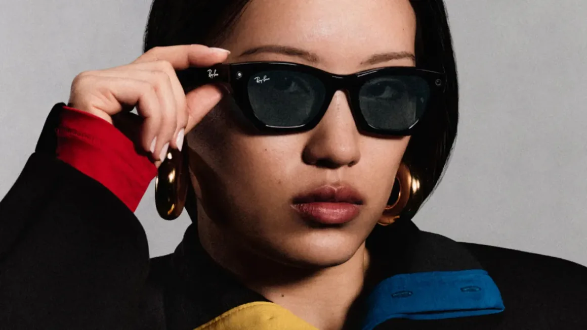 Koyu saçlı bir kişi siyah Ray-Ban güneş gözlüğü ve büyük altın halka küpeler takıyor; eli gözlüğün çerçevesinde.
