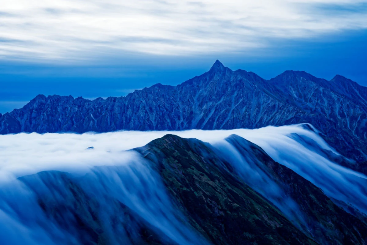 74. Nikkor Photo Contest Kazananları Açıklandı 28 A long exposure shot captures a sea of clouds flowing over dark, jagged mountain ridges under a deep blue twilight sky.
