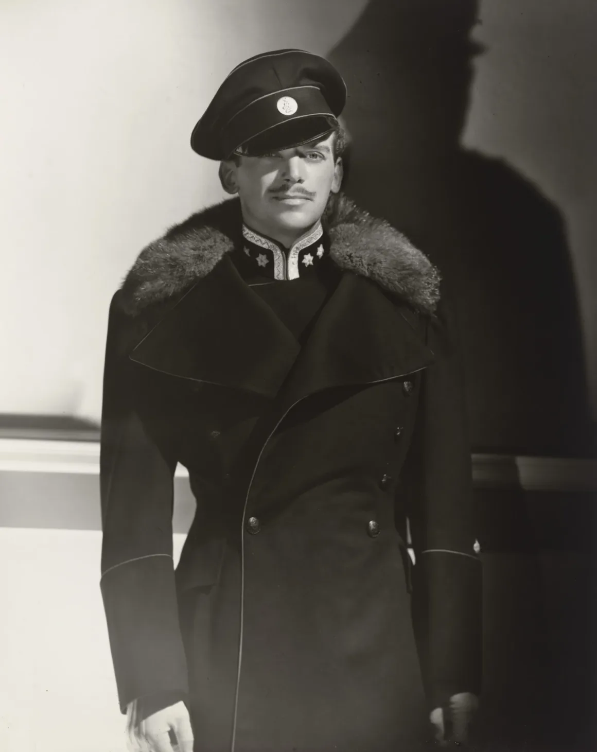 Hollywood’un Altın Çağındaki Glamoru Fotoğraflayan Adam: George Hurrell 26 İnce bıyıklı bir adam, kürk yakalı koyu renk askeri tarzda palto ve üzerinde yuvarlak arma bulunan siperlikli şapka giyiyor.