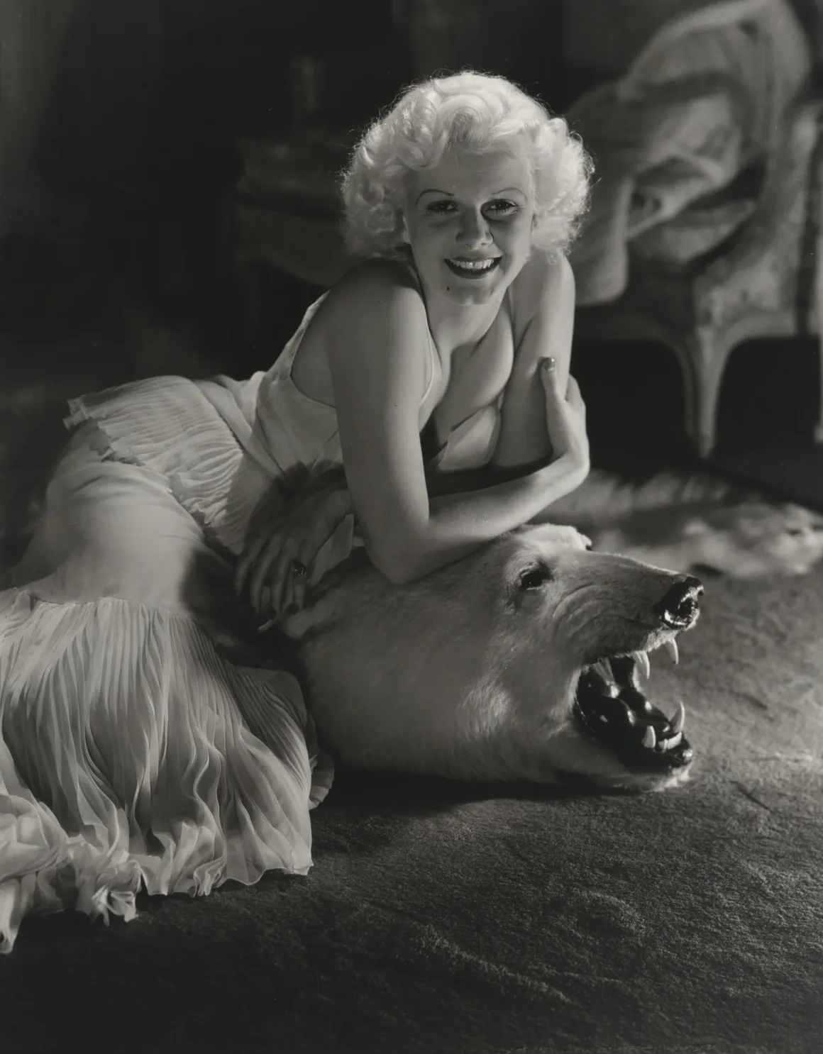 Hollywood’un Altın Çağındaki Glamoru Fotoğraflayan Adam: George Hurrell 24 Sarı saçlı bir kadın, açık renkli pileli elbisesiyle gülümserken kollarını tahnit edilmiş kutup ayısı postunun başına yaslıyor.