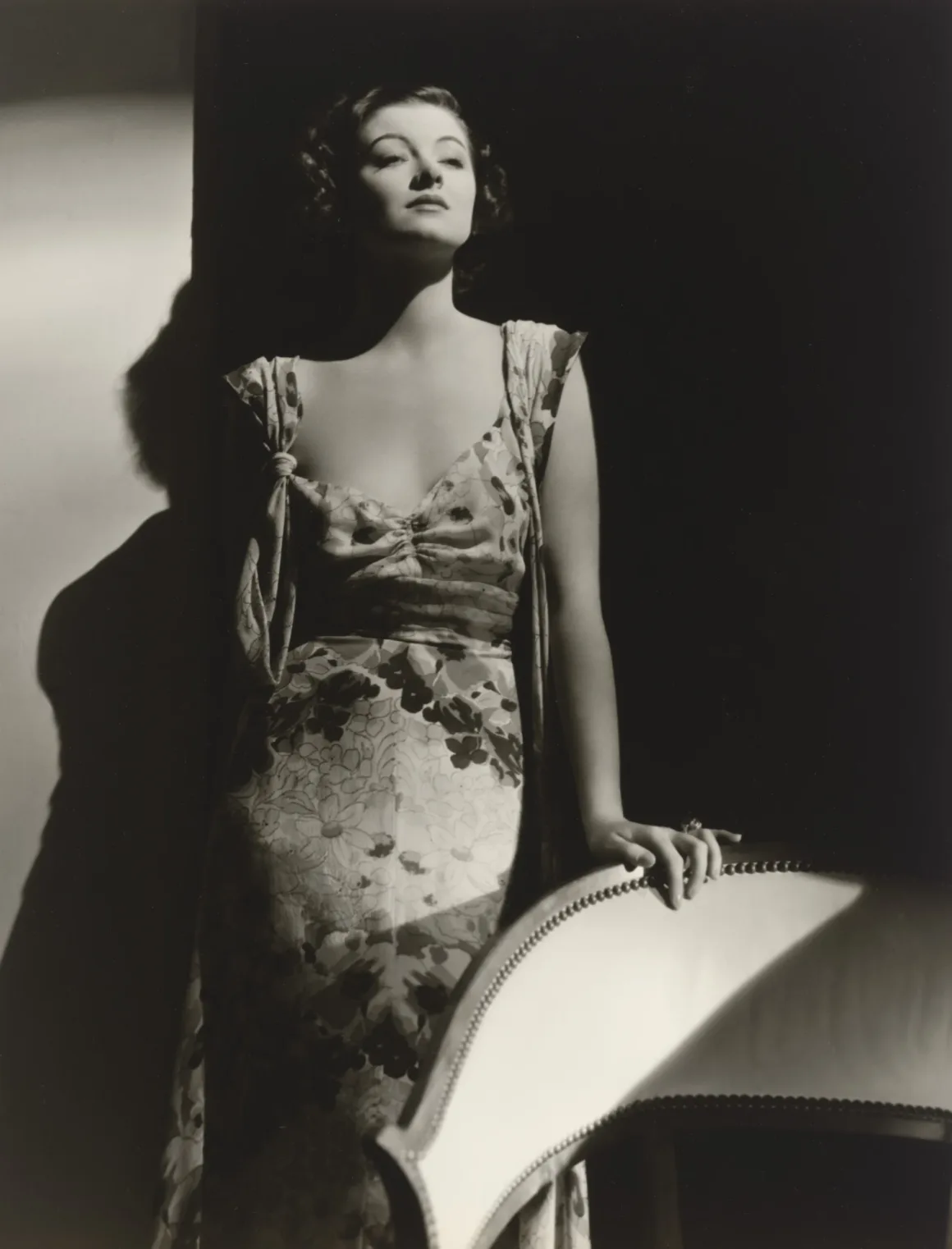 Hollywood’un Altın Çağındaki Glamoru Fotoğraflayan Adam: George Hurrell 21 Koyu dalgalı saçlı bir kadın, çiçek desenli elbisesiyle bir sandalyeye yaslanarak yüksek kontrastlı siyah-beyaz portre için poz veriyor.
