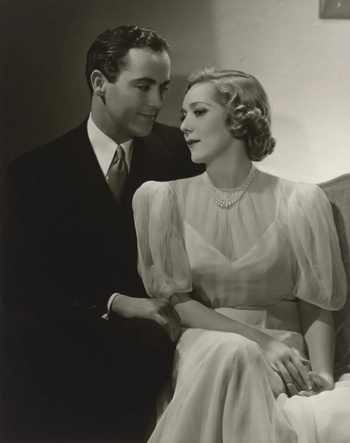Hollywood’un Altın Çağındaki Glamoru Fotoğraflayan Adam: George Hurrell 25 Koyu renk takım elbise ve kravat giyen bir adam, açık renk kısa kollu elbise ve inci kolye takan bir kadının yanında oturuyor.