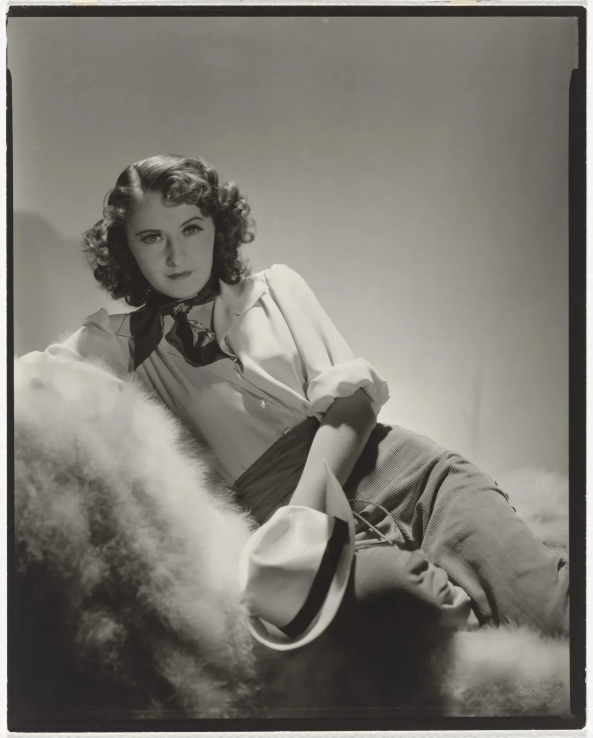 Hollywood’un Altın Çağındaki Glamoru Fotoğraflayan Adam: George Hurrell 23 Dalgalı kahverengi saçlı bir kadın, beyaz yakalı gömlek, koyu renk boyun bağı ve açık renk pantolonla kürk kaplı bir yüzeye uzanmış, elinde beyaz bir fötr şapka tutuyor.