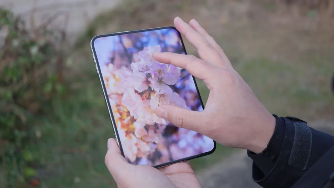 iPhone Duo İncelemesi: Etkileyici Tasarım, Kaçınılmaz Tavizler 24 Bir kişi, ekranda pembe kiraz çiçeklerinin yakın plan fotoğrafını gösteren katlanabilir akıllı telefonu parmaklarıyla kullanıyor.