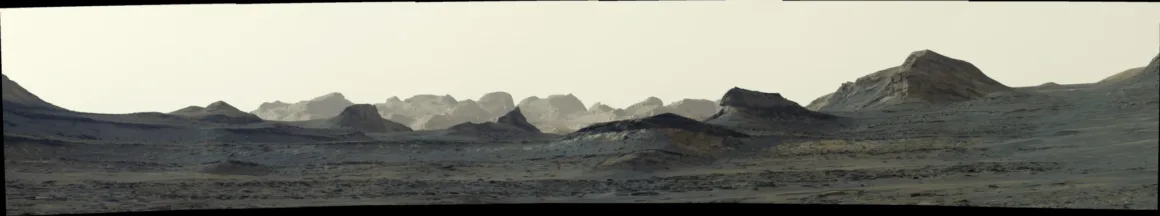 Curiosity’den Bir Çarpıcı Mars Karesi Daha: Gün Doğumunda Sharp Dağı 19 Soluk ve puslu bir gökyüzünün altında katmanlı tepeler ile uzaktaki dağların görüldüğü geniş, kayalık Mars manzarası.