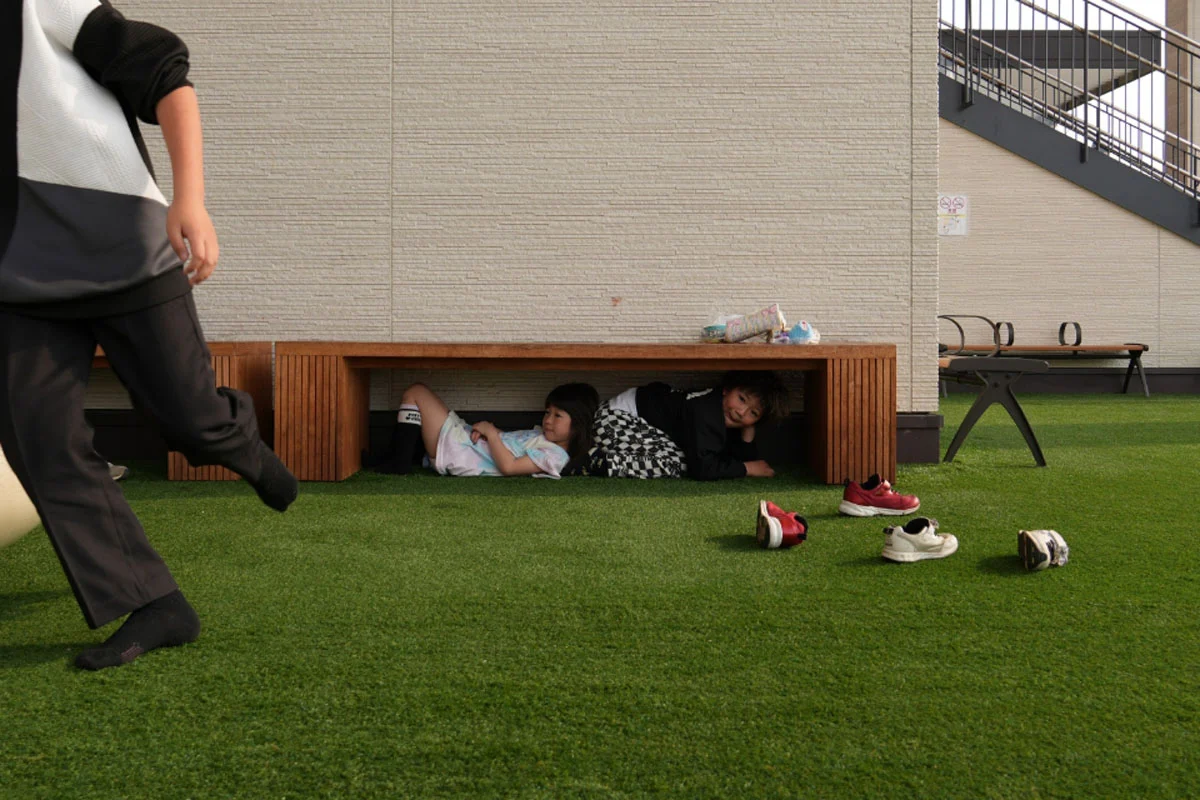 74. Nikkor Photo Contest Kazananları Açıklandı 49 Two children lie underneath a wooden bench on a grassy outdoor area, while a person walks past in the foreground and several shoes are scattered on the grass.