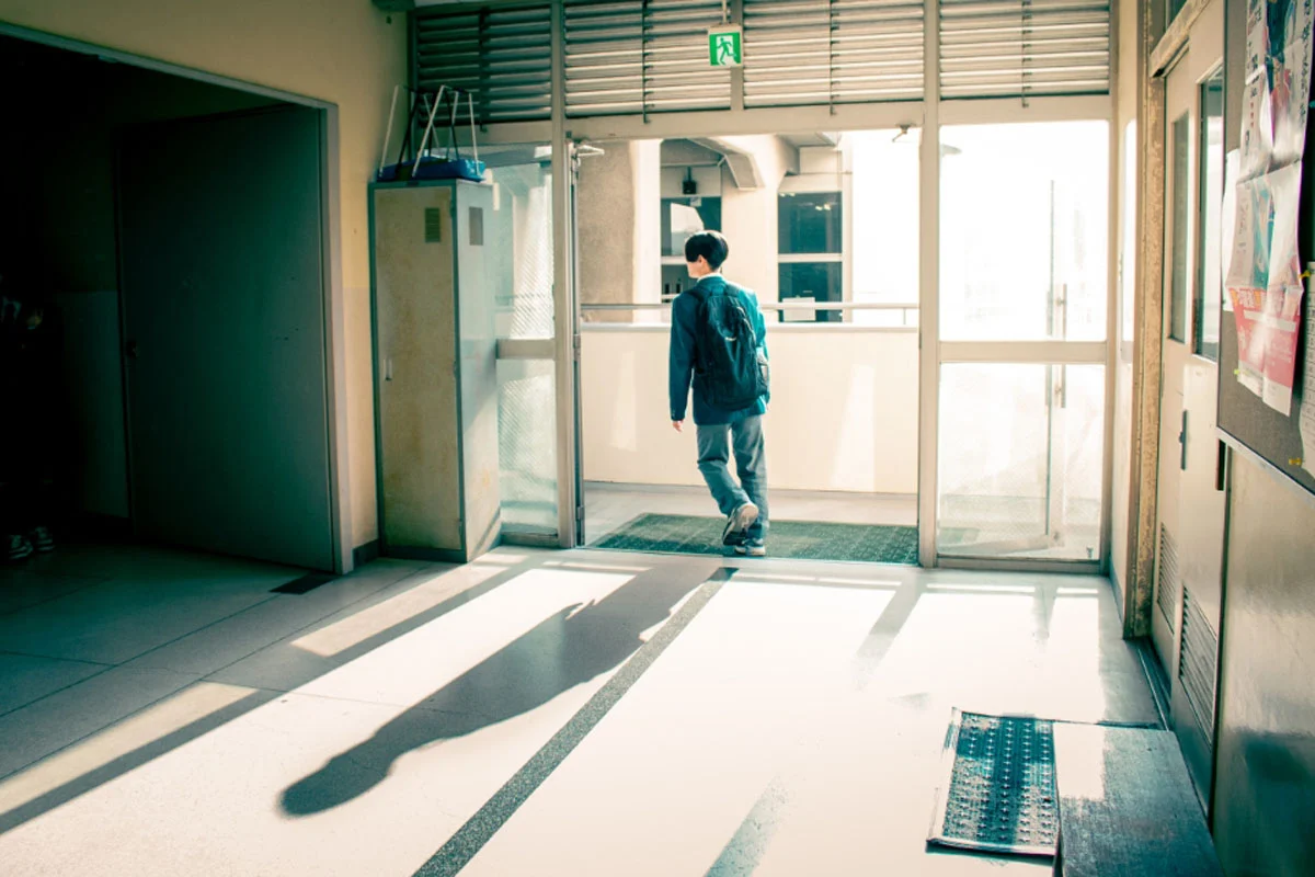 74. Nikkor Photo Contest Kazananları Açıklandı 47 A student with a backpack walks through an open doorway into a bright, sunlit corridor, casting a long shadow on the floor.