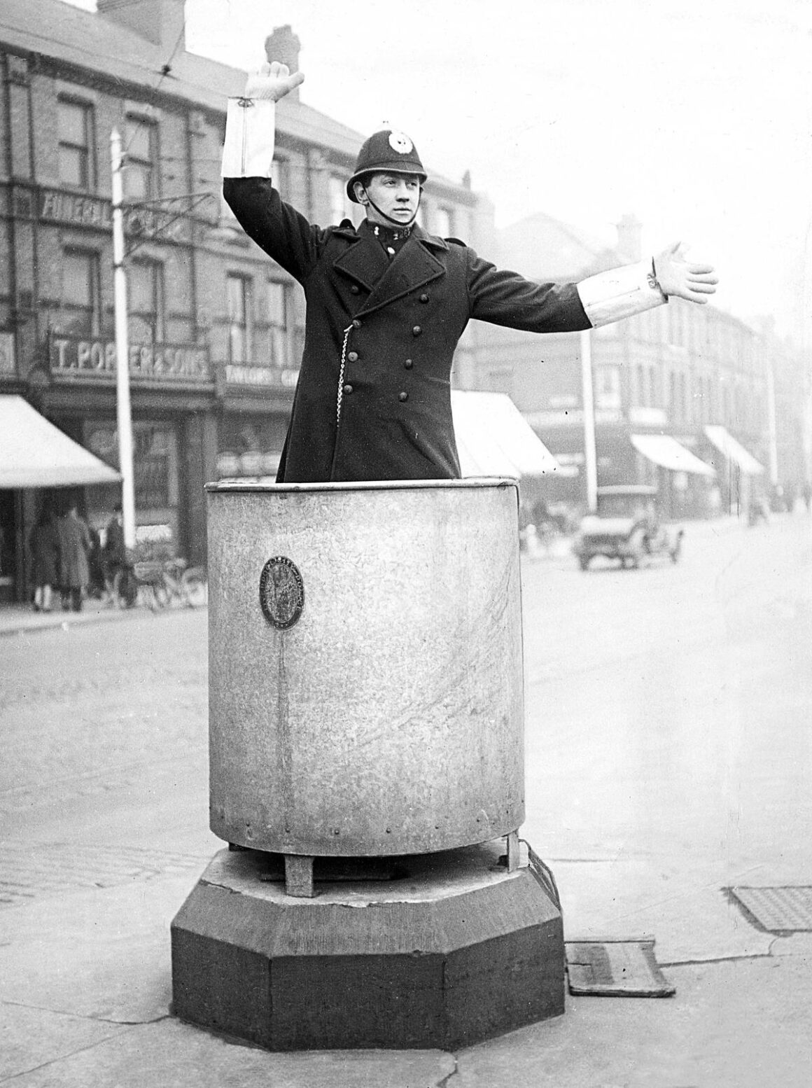 1930’ların Britanyası: Gündelik Hayatı Belgeleyen Unutulmuş Fotoğraflar 16 Miğfer ve uzun palto giyen bir polis memuru, Liverpool'da bir caddenin ortasındaki kaide üzerine yerleştirilmiş büyük metal bir silindirin içinde duruyor; kollarını açarak trafiği yönlendiriyor.