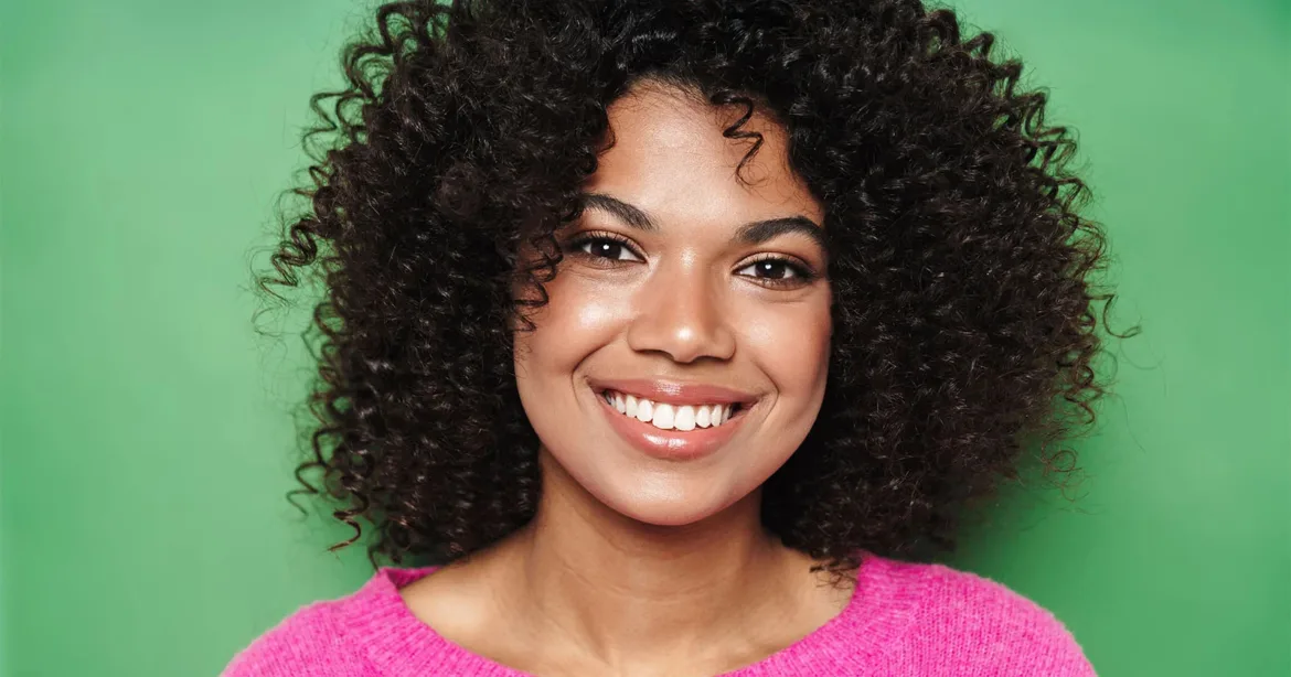 A smiling woman with dark, curly hair wears a bright pink sweater against a solid green background.