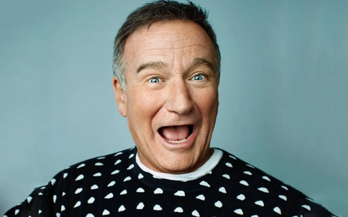 Fotoğraflarda Neden Gülümsüyoruz? Portre Geleneğinin Değişen Yüzü 21 Robin Williams düz mavi arka plan önünde ağzı açık biçimde genişçe gülümsüyor ve beyaz bulut desenli siyah bir kazak giyiyor.