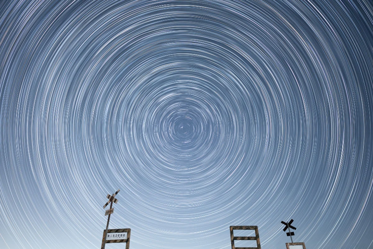 74. Nikkor Photo Contest Kazananları Açıklandı 34 A long-exposure star trail photograph showing concentric circles of light in the night sky above two railway crossing signs.
