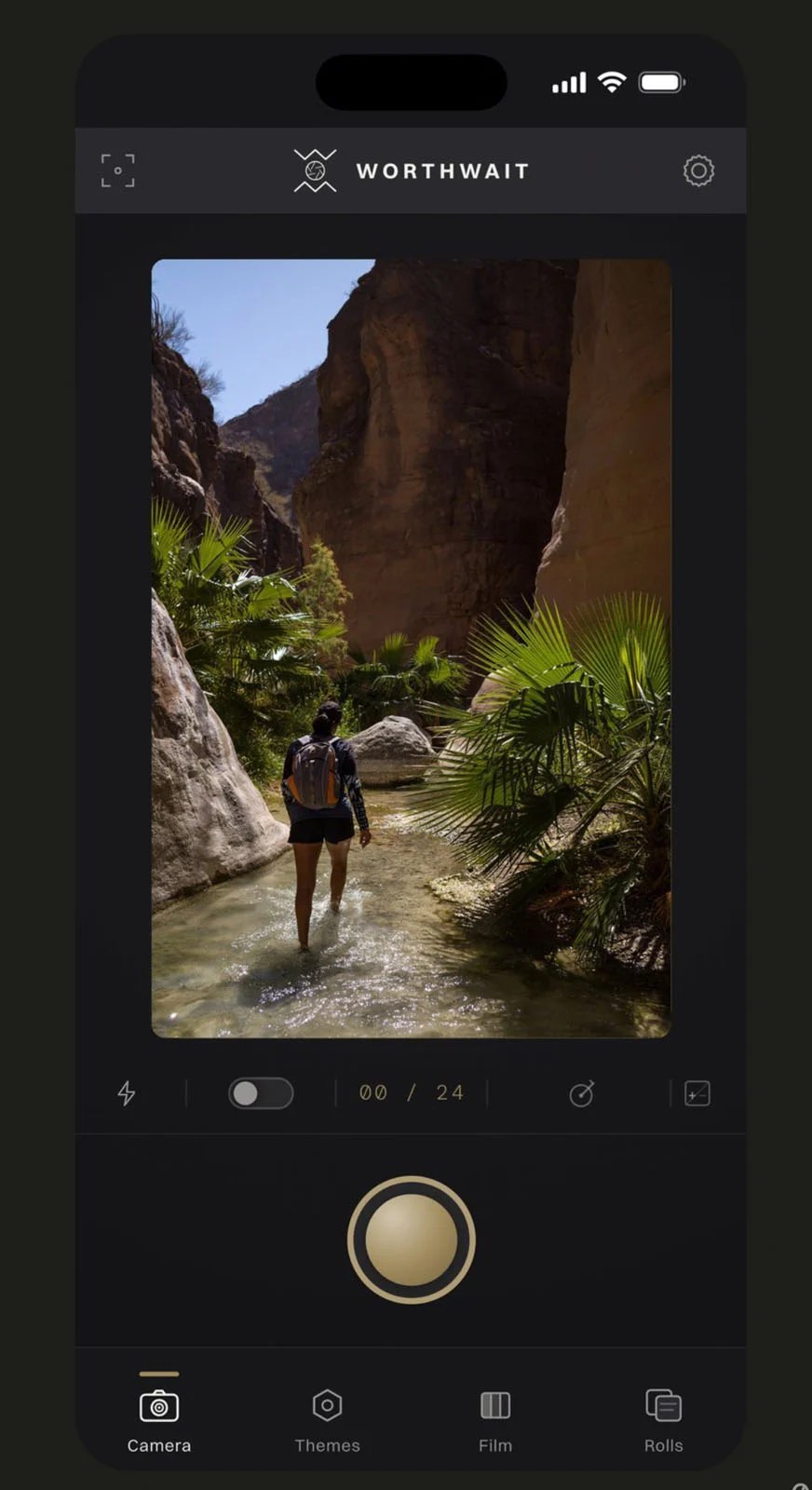 WorthWait: Fotoğrafları Görmek İçin 24 Saat Bekleten Kamera Uygulaması 24 Akıllı telefon ekranında WorthWait kamera uygulaması açık; kadrajda bir kanyon içindeki sığ derede yürüyen kişi görülüyor.