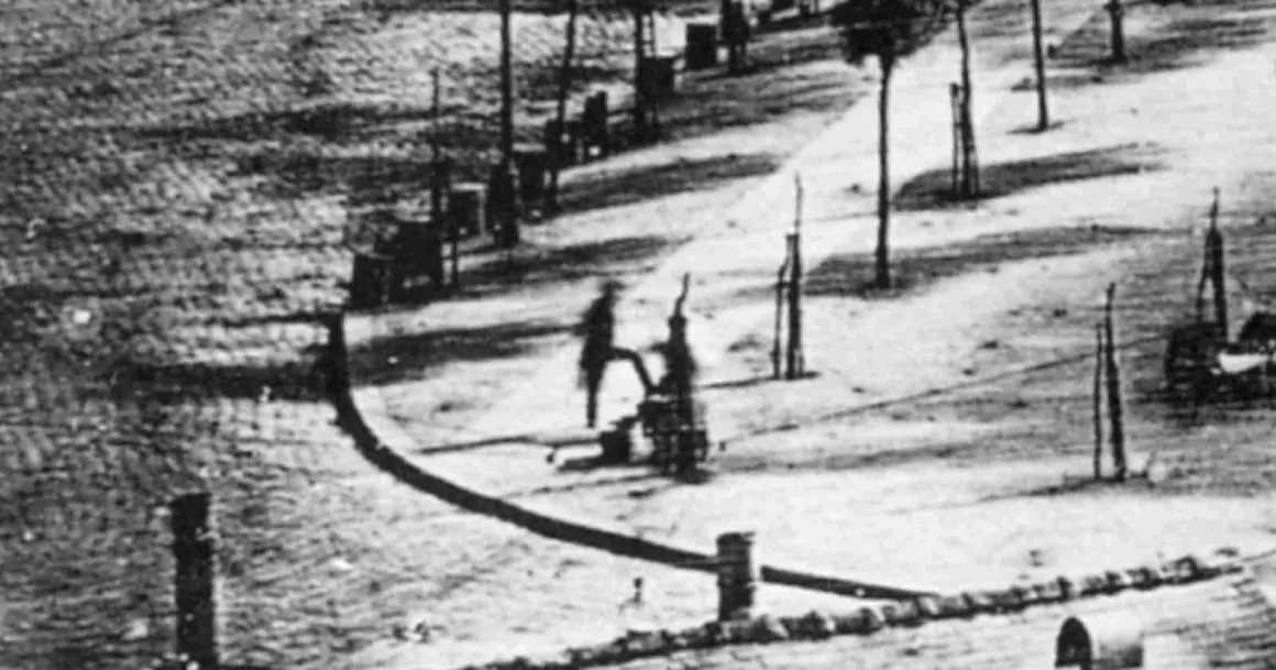 Bulanık siyah-beyaz uzun pozlama fotoğrafında, kaldırımda ayakkabıları boyanırken duran bir kişi görülüyor.